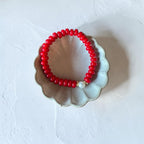 A red beaded bracelet with a small white pearl accent, displayed on a scalloped ceramic dish against a textured white background.