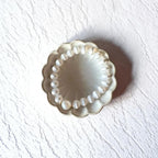 A scalloped cream-colored ceramic dish or jewelry holder containing a white pearl beaded bracelet, set against a textured white background with a soft shadow.