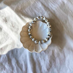 A beaded stretch bracelet made of white beads with black speckles, displayed on a small scalloped cream dish over a textured white fabric background.
