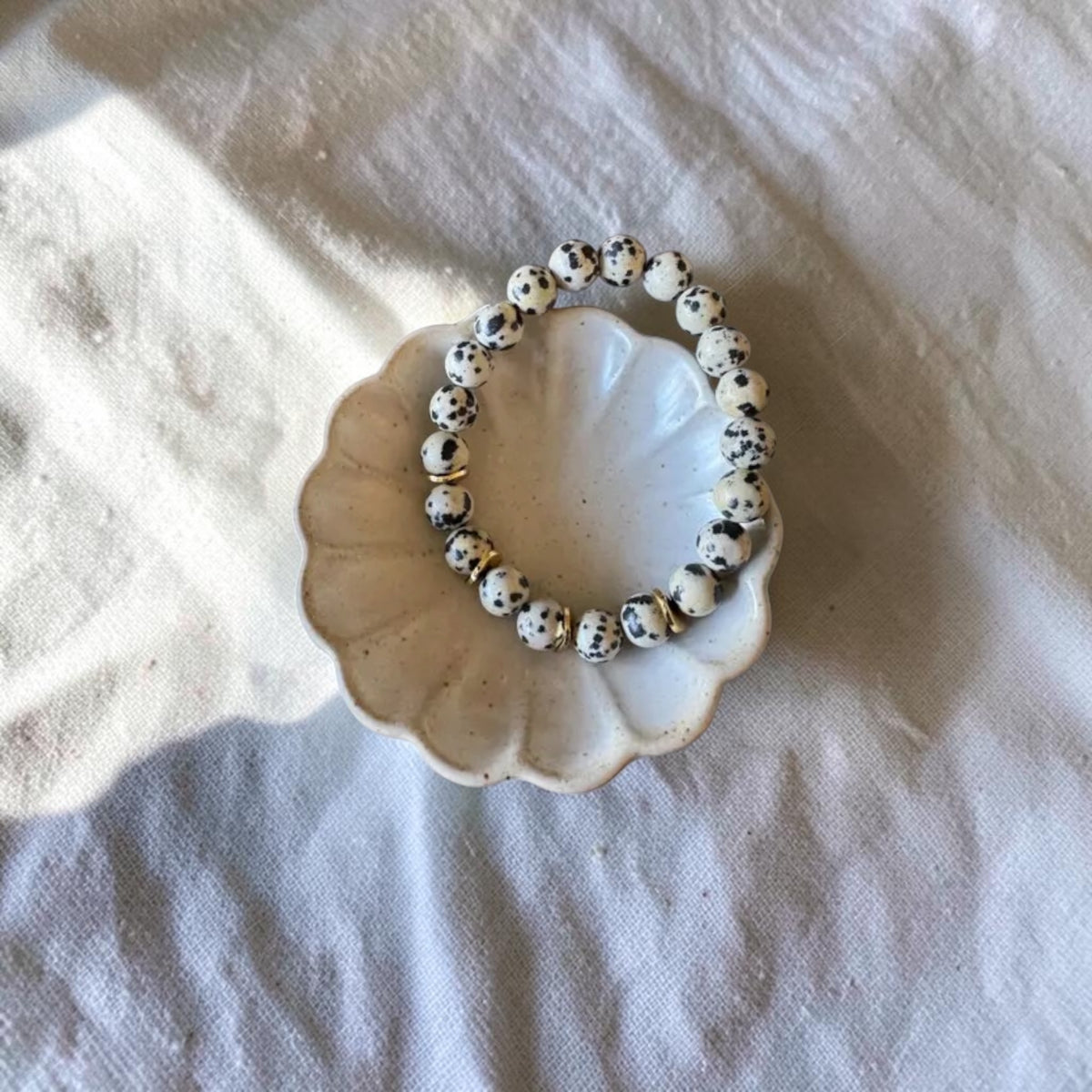 A beaded stretch bracelet made of white beads with black speckles, displayed on a small scalloped cream dish over a textured white fabric background.