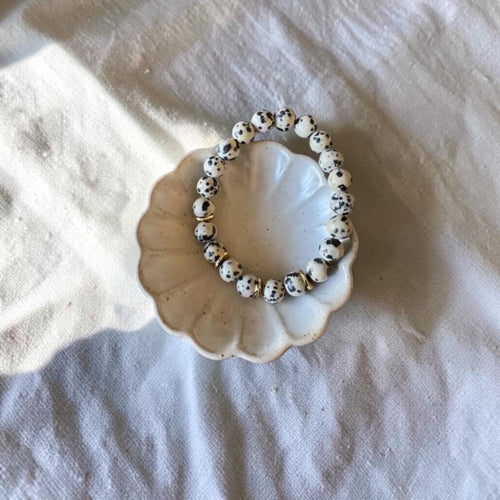 A beaded stretch bracelet made of white beads with black speckles, displayed on a small scalloped cream dish over a textured white fabric background.