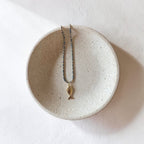 A delicate necklace with a small gold fish pendant and a dark beaded chain, displayed on a round, speckled ceramic dish against a textured white background.
