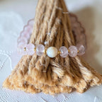 A beaded bracelet with light pink rose quartz beads and a central white pearl, displayed on a rustic jute cone stand.