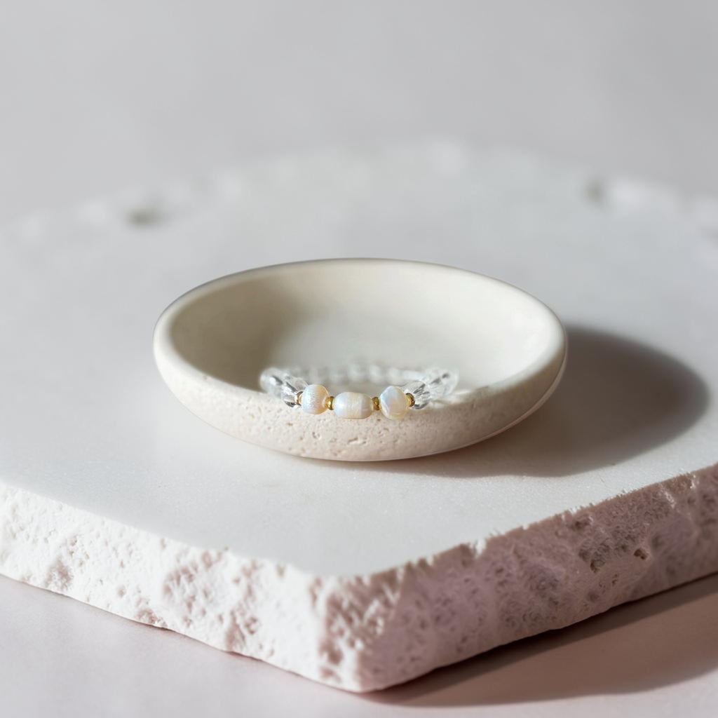 A delicate silver-toned beaded bracelet with small white pearls and gold accents, displayed in a small round ceramic dish on a textured stone slab.