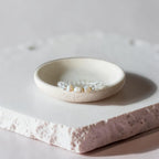 A delicate silver-toned beaded bracelet with small white pearls and gold accents, displayed in a small round ceramic dish on a textured stone slab.