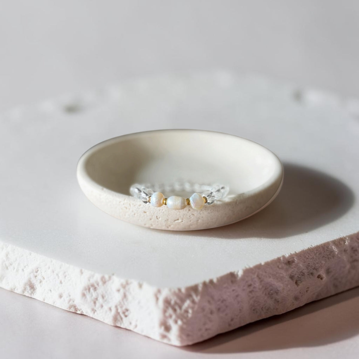 A delicate silver-toned beaded bracelet with small white pearls and gold accents, displayed in a small round ceramic dish on a textured stone slab.