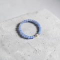 A light blue beaded stretch bracelet with a small silver heart charm, resting on a textured light gray concrete block.