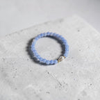 A light blue beaded stretch bracelet with a small silver heart charm, resting on a textured light gray concrete block.