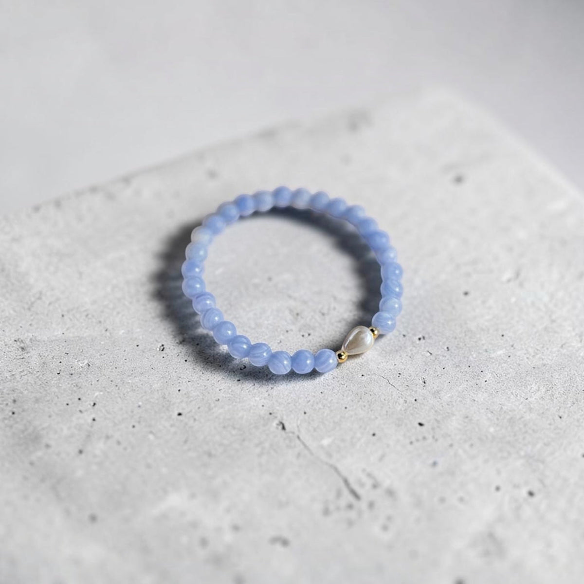 A light blue beaded stretch bracelet with a small silver heart charm, resting on a textured light gray concrete block.