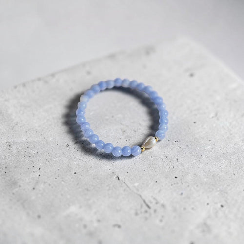 A light blue beaded stretch bracelet with a small silver heart charm, resting on a textured light gray concrete block.