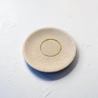 A round, shallow ceramic dish in a speckled beige finish with a thin, hand-painted green ring detail in the center, set against a textured white background.