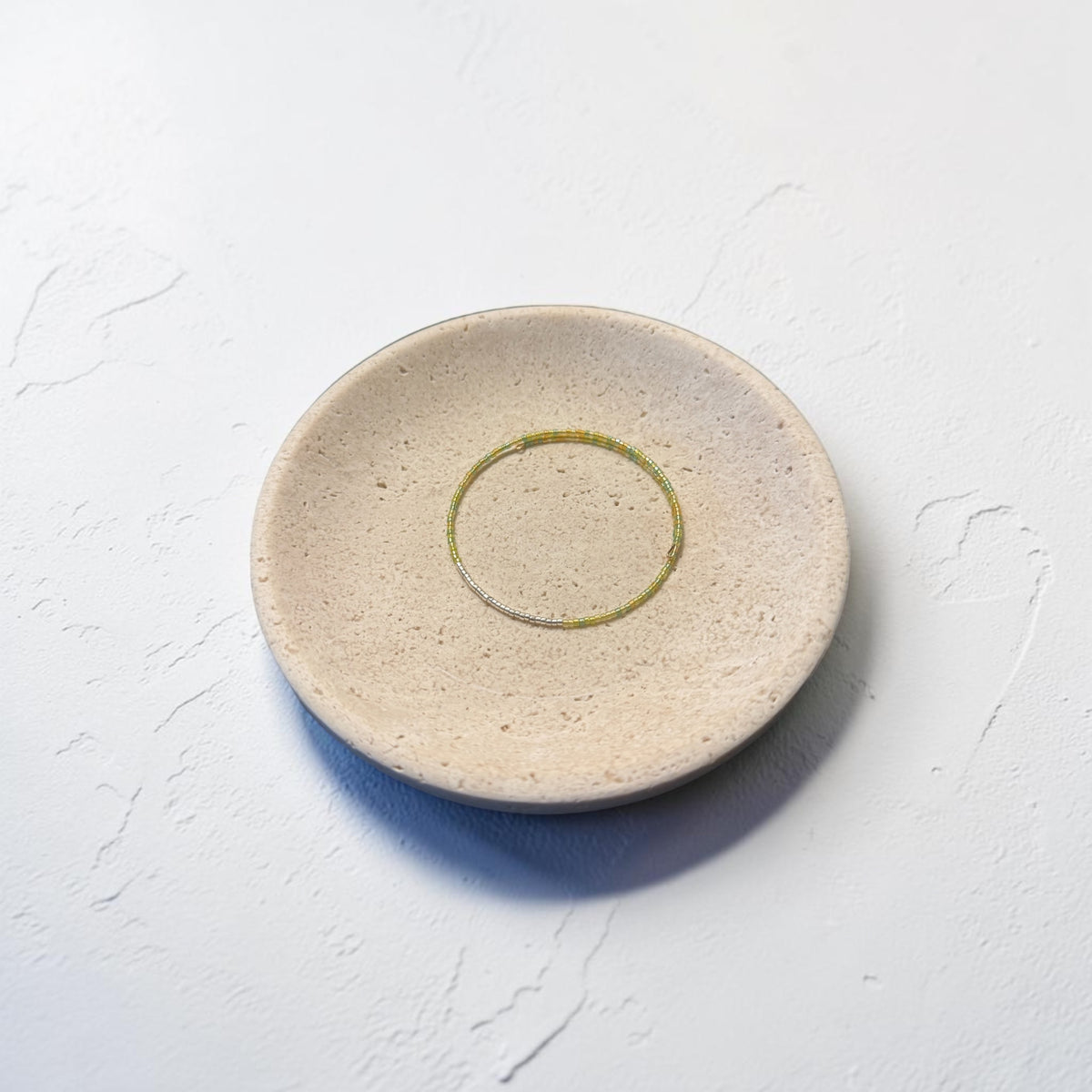 A round, shallow ceramic dish in a speckled beige finish with a thin, hand-painted green ring detail in the center, set against a textured white background.