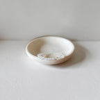 A delicate clear beaded bracelet with a small long pearl charm, displayed inside a small, shallow, textured cream ceramic dish on a neutral light gray surface.