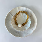 A translucent amber-yellow beaded bracelet with a white pearl-like bead, displayed on a white ceramic flower-shaped dish inside a larger speckled off-white plate against a light gray background.