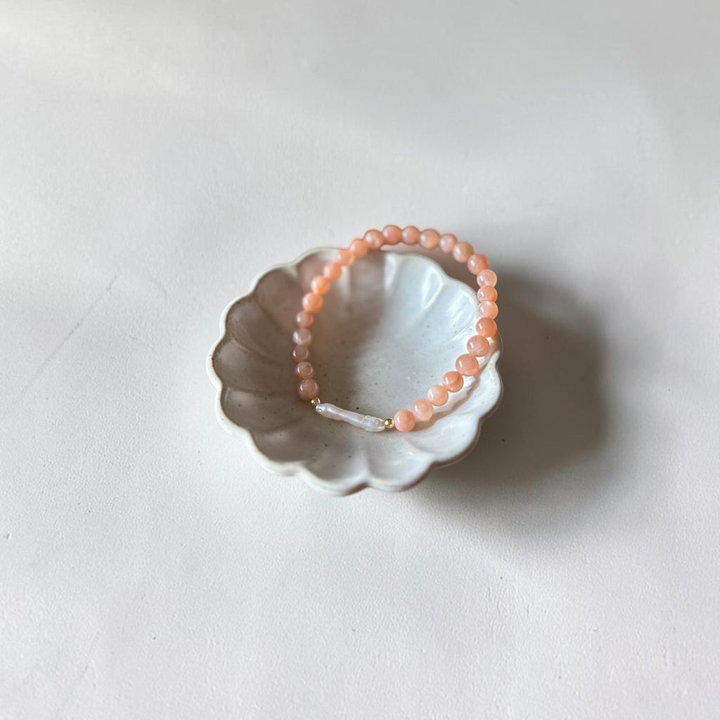 A peach-colored beaded bracelet with a small white bead and gold clasp, displayed on a small white ceramic dish against a light gray background.