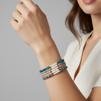 A close-up of a woman's wrist wearing a stack of four beaded bracelets in teal, cream, and slate blue, against a soft gray studio background.