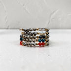 A stack of beaded stretch bracelets displayed on a white surface against a textured white wall. The bracelets feature speckled gray and black beads, white beads, gold spacer beads, and small accent beads in red, teal blue, and white.