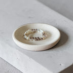A beaded stretch bracelet with white pearls and brown beads resting on a small round ceramic dish, set on a light gray concrete block.