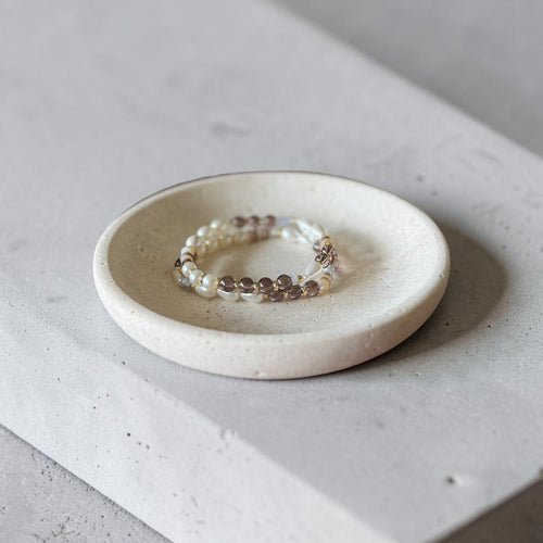 A beaded stretch bracelet with white pearls and brown beads resting on a small round ceramic dish, set on a light gray concrete block.