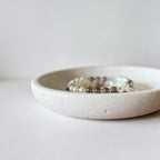 A beaded stretch bracelet with white, brown, and clear beads resting inside a small, round, textured off-white ceramic dish on a light gray surface.