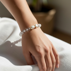A close-up of a person's wrist wearing a beaded bracelet made of clear faceted beads and white pearl-like beads, resting on a white fabric surface.