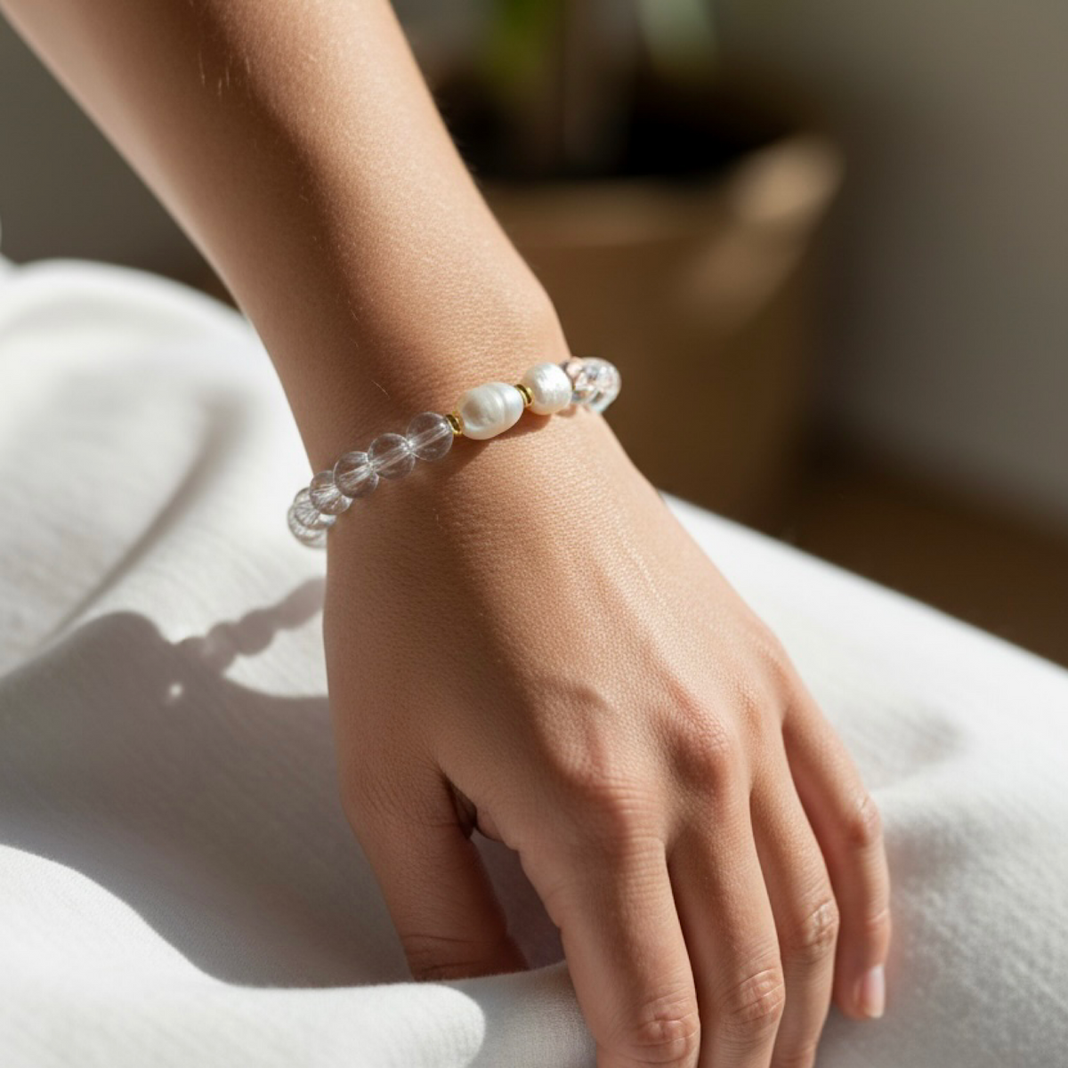A close-up of a person's wrist wearing a beaded bracelet made of clear faceted beads and white pearl-like beads, resting on a white fabric surface.