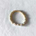 A gold beaded stretch bracelet with three white textured glass beads, displayed on a textured white surface.
