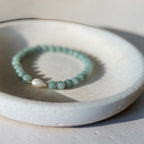 A beaded stretch bracelet with light green stone beads and a white teardrop charm, resting on a textured white ceramic dish.