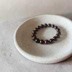 A beaded stretch bracelet made of dark brown polished round beads, displayed on a small textured off-white ceramic dish against a neutral gray background.
