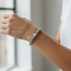 A close-up of a person's wrist wearing a beaded bracelet with light green and white beads, set against a soft-focus indoor background near a window.
