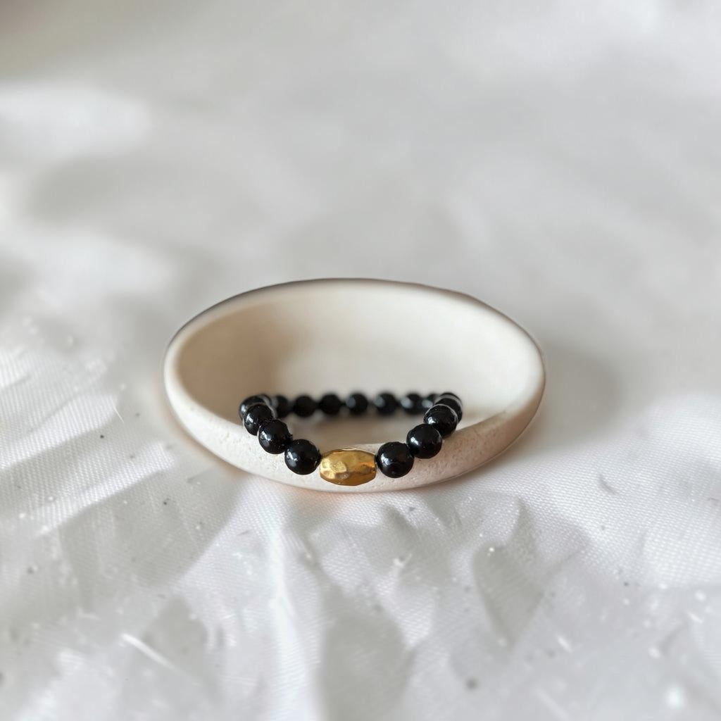 A beaded stretch bracelet with black round beads and a single gold oval accent bead, displayed on a small white ceramic dish over a white textured fabric background.