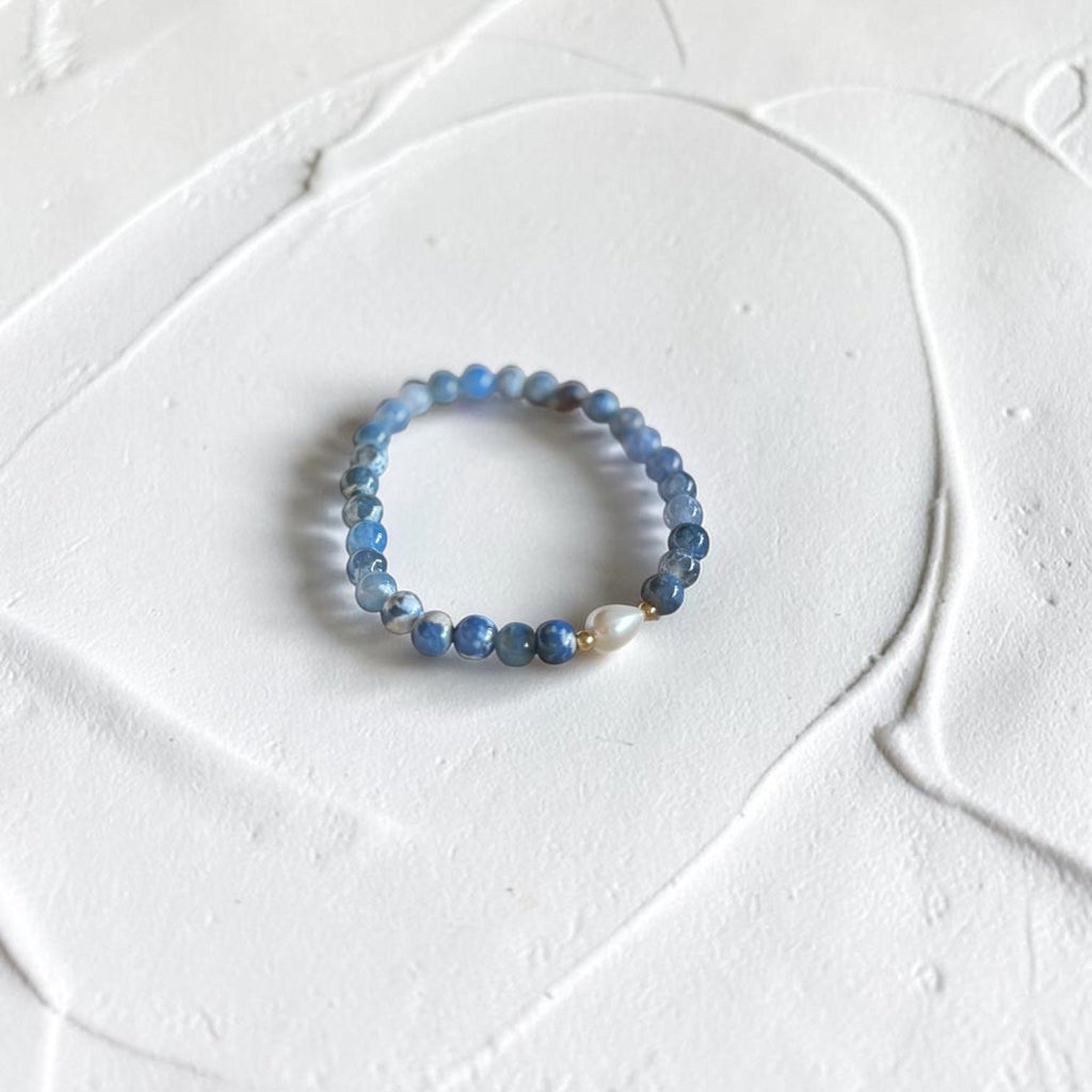 A beaded stretch bracelet featuring light blue and dark blue round beads with a single white teardrop bead, displayed on a textured white surface.