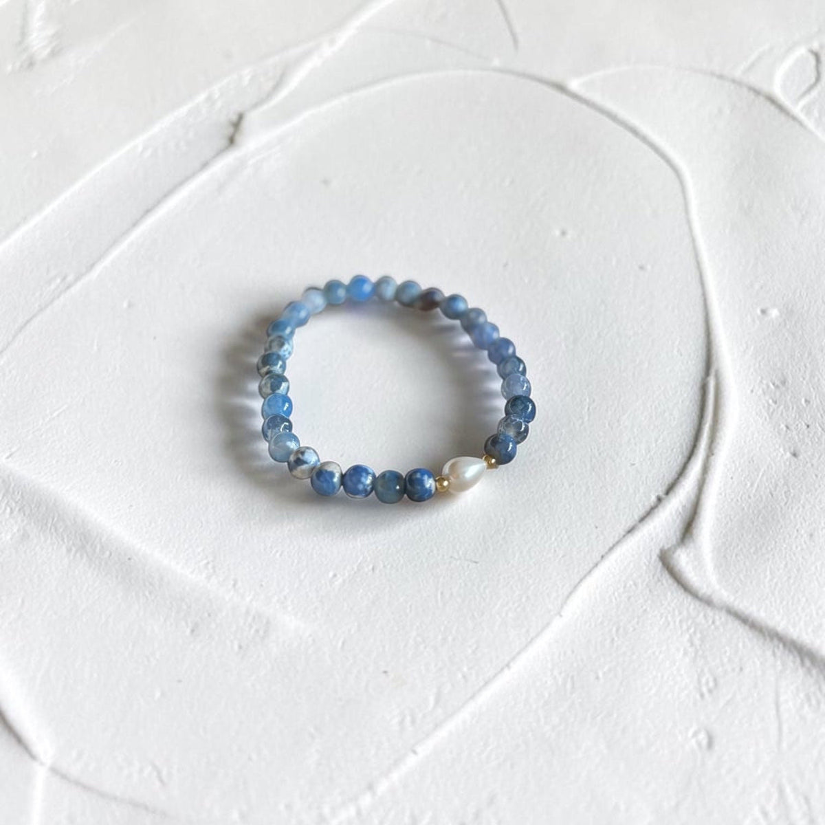 A beaded stretch bracelet featuring light blue and dark blue round beads with a single white teardrop bead, displayed on a textured white surface.