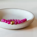 A beaded bracelet with bright pink round beads and a single central gold metallic bead, displayed on a white ceramic dish against a light beige background.