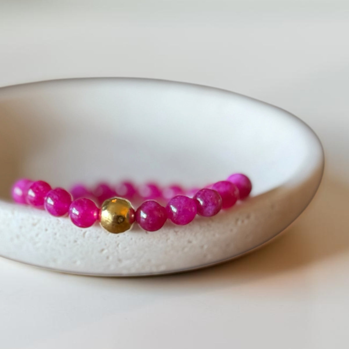 A beaded bracelet with bright pink round beads and a single central gold metallic bead, displayed on a white ceramic dish against a light beige background.