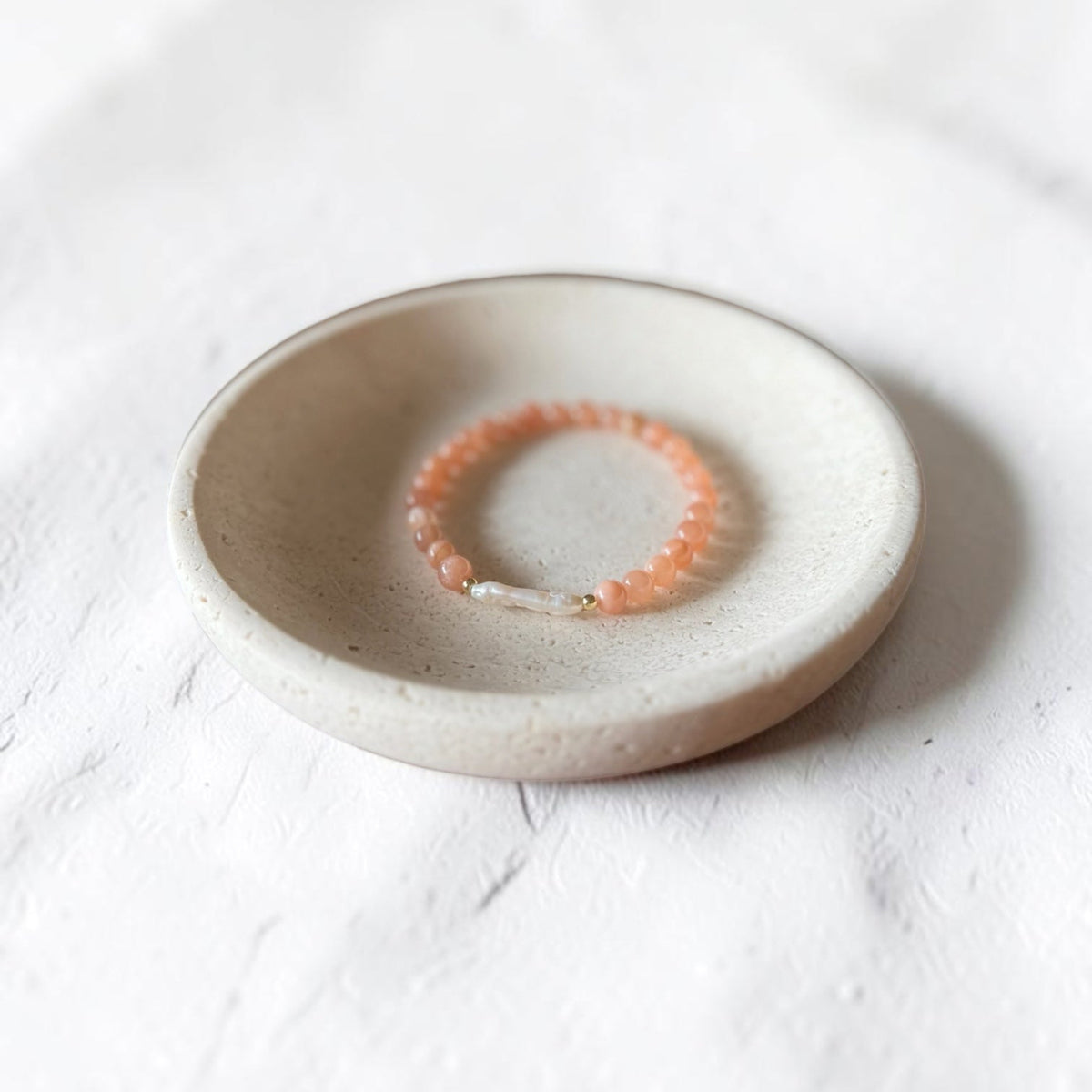 A peach-orange beaded bracelet with a small gold charm, displayed inside a round, speckled beige ceramic dish on a white textured surface.