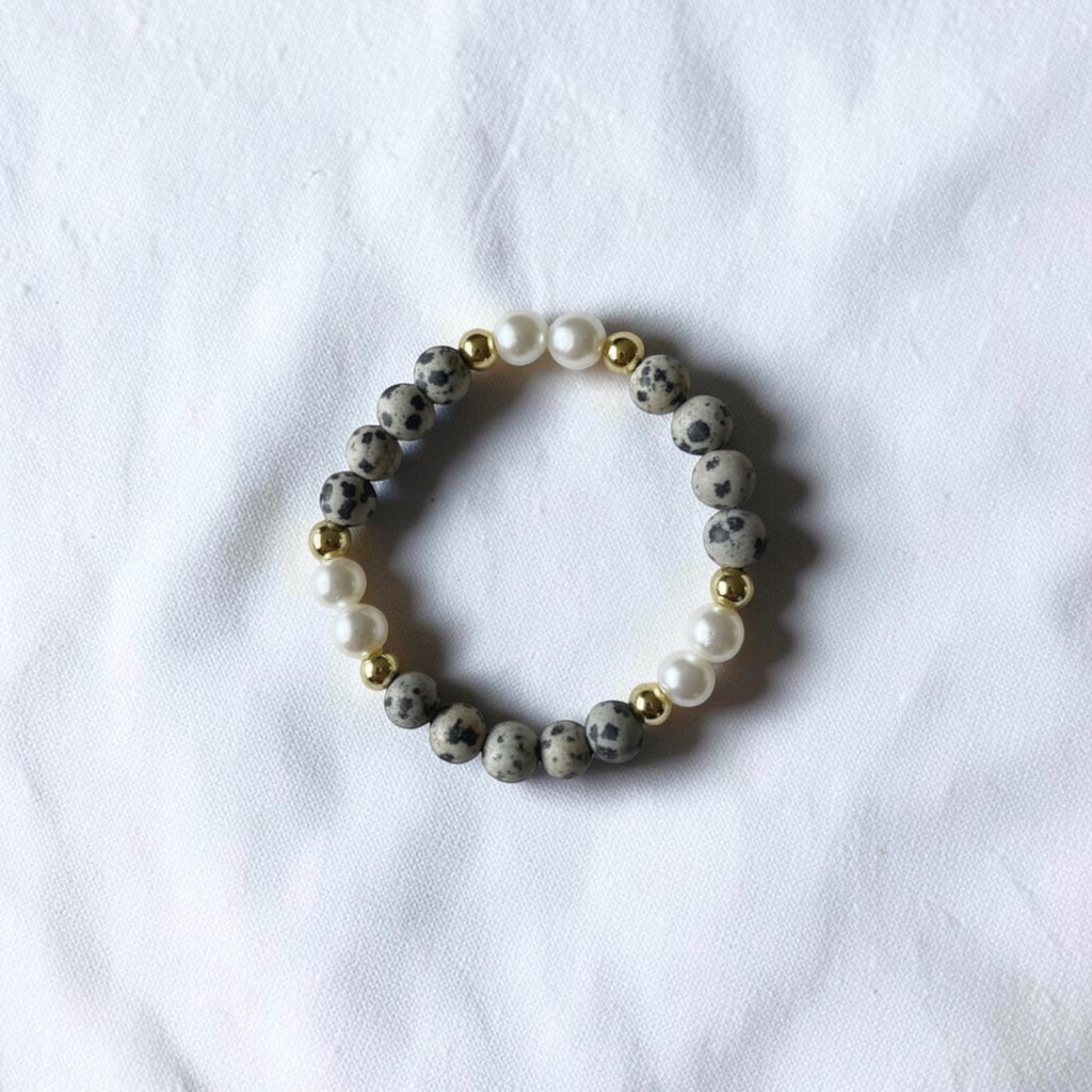 A beaded stretch bracelet featuring gray and black speckled stone beads, white faux pearls, and small gold spacer beads, displayed on a textured white fabric background.