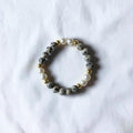A beaded stretch bracelet featuring gray and black speckled stone beads, white faux pearls, and small gold spacer beads, displayed on a textured white fabric background.