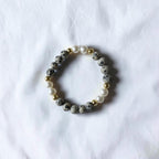 A beaded stretch bracelet featuring gray and black speckled stone beads, white faux pearls, and small gold spacer beads, displayed on a textured white fabric background.