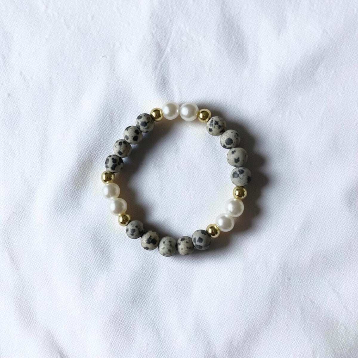 A beaded stretch bracelet featuring gray and black speckled stone beads, white faux pearls, and small gold spacer beads, displayed on a textured white fabric background.
