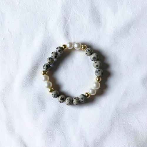 A beaded stretch bracelet featuring gray and black speckled stone beads, white faux pearls, and small gold spacer beads, displayed on a textured white fabric background.