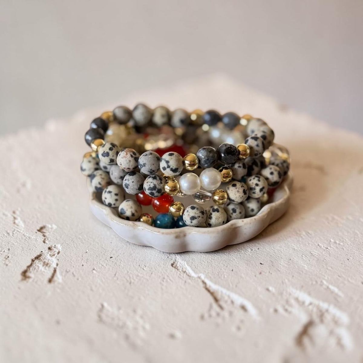 A stack of beaded stretch bracelets displayed on a small white ceramic dish. The bracelets feature white and black speckled dalmatian-style beads, dark gray beads, white pearl-like beads, and small metallic gold and red accent beads.