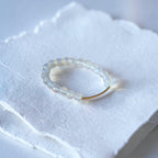A delicate beaded ring with pale blue-gray translucent beads and a single gold-toned bead, resting on a textured white paper surface.