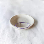 A thin purple beaded bracelet displayed inside a small, round, speckled off-white ceramic dish on a textured white surface.