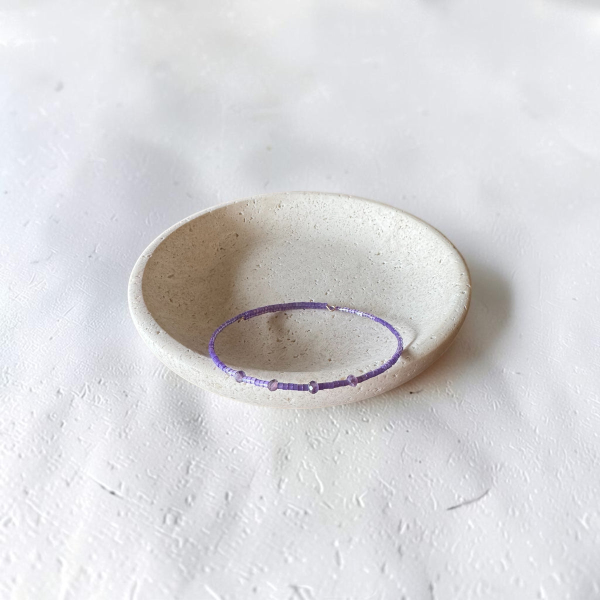 A thin purple beaded bracelet displayed inside a small, round, speckled off-white ceramic dish on a textured white surface.