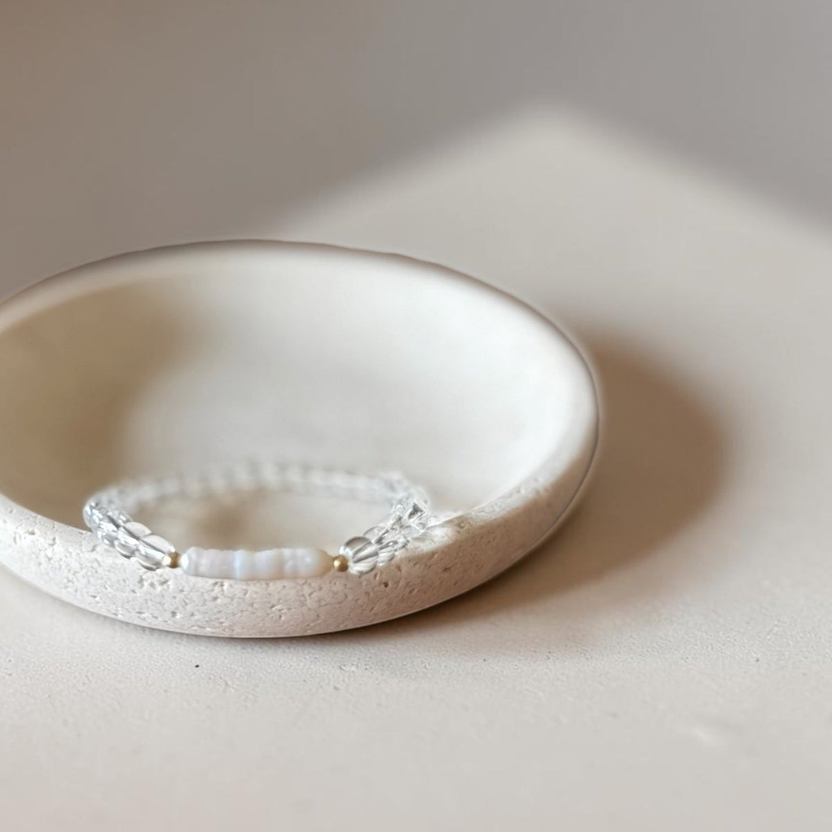 A delicate clear beaded bracelet with pearl and gold filled accents resting inside a small, textured off-white ceramic dish on a neutral beige surface.