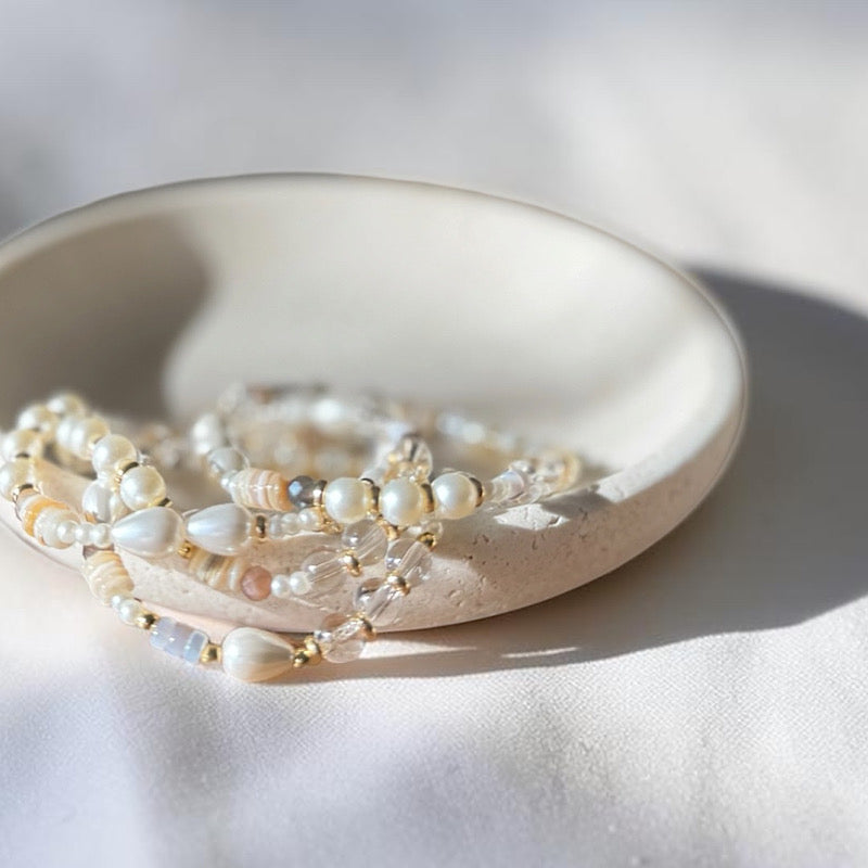 A close-up of delicate beaded bracelets with white pearls, cream beads, and small gold-toned accents, arranged in a shallow round ceramic dish on a softly lit white fabric surface.