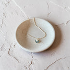 A delicate necklace with a small blue and white fan-shaped pendant resting on a round, textured off-white ceramic dish, set against a matching textured light pink background.