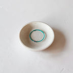 A thin turquoise metal bangle bracelet displayed inside a small, round, speckled off-white ceramic dish on a textured light gray surface.