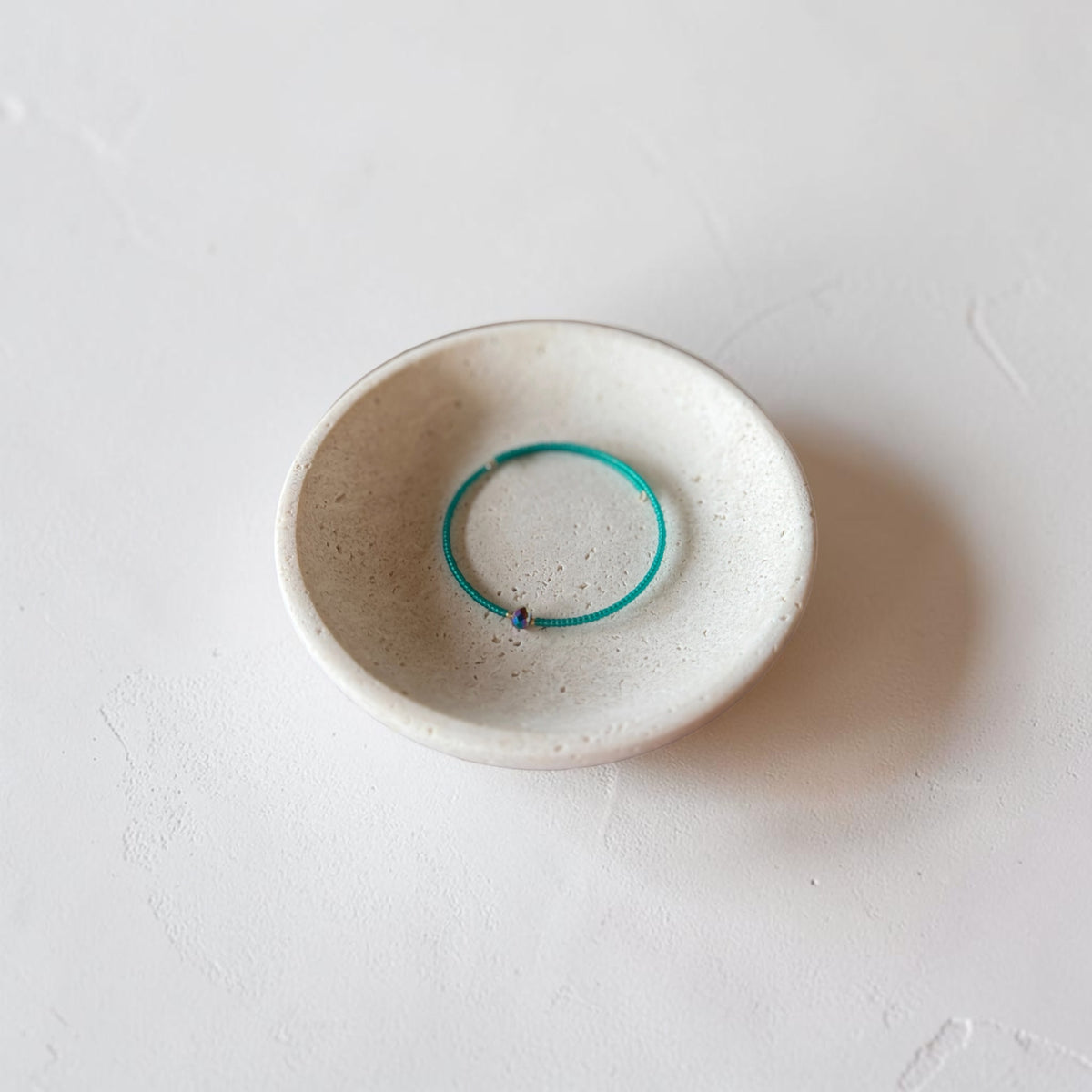 A thin turquoise metal bangle bracelet displayed inside a small, round, speckled off-white ceramic dish on a textured light gray surface.